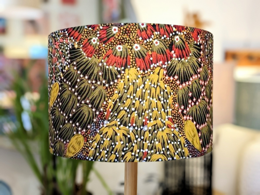 The Bush Banana Lampshade by Artist Donna Abbot displays an intricate abstract pattern of red, yellow, black, and white dots and lines inspired by nature and Indigenous art, set against a soft indoor backdrop.