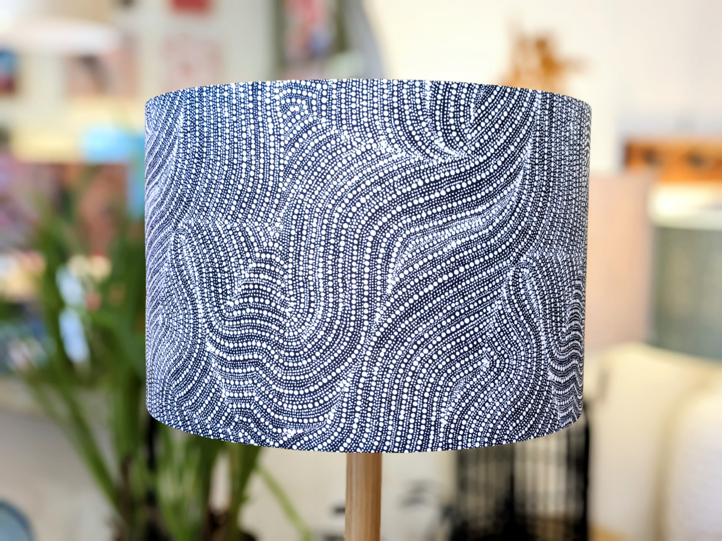 The Desert Dreaming Aboriginal lampshade by artist Senita Napangardi Granites showcases a handmade blue and white abstract, wavy dotted pattern inspired by Desert Fringe-Rush Seed Dreaming, with indoor plants and furniture softly blurred in the background.