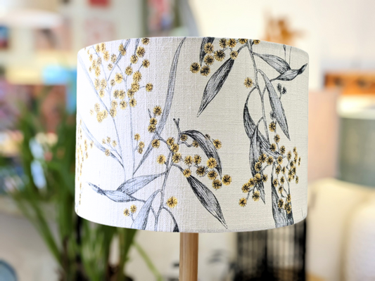A close-up of the Emotive Collections Golden Wattle Lampshade, showcasing a drum style with yellow wattle flowers and gray leaves on cotton slub fabric, set indoors with softly blurred decor in the background.