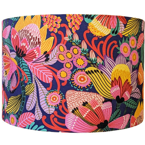The Wild Protea Blue Lampshade from the Emotive Collection features a vibrant floral pattern with bold pink, yellow, orange, red, and green flowers and leaves on a dark blue background—designed by Kirsten Katz.