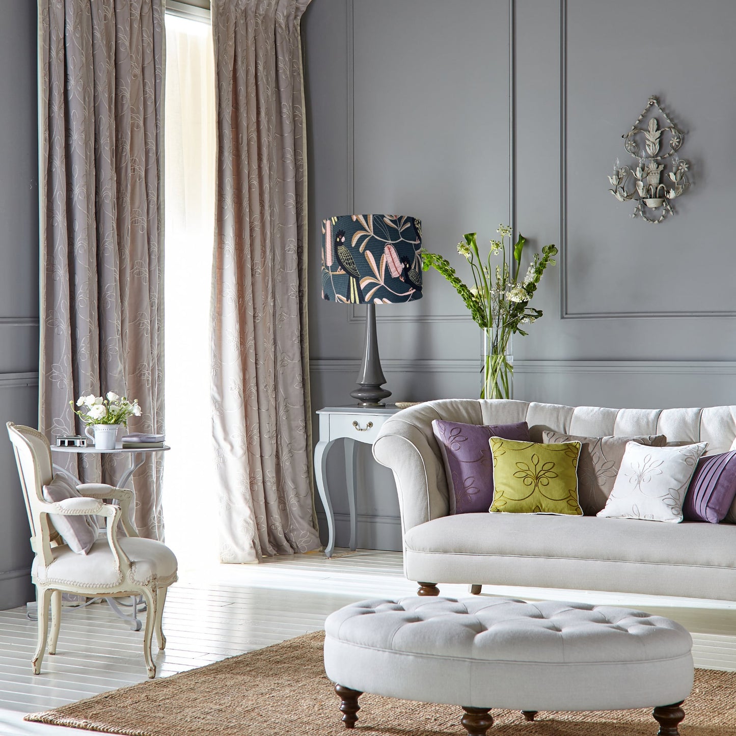 Elegant living room with grey walls, white sofa with colorful pillows from the Jocelyn Proust Australian Flora Collection, round tufted ottoman, floral vase display, tall curtains, and an Emotive Collection Yellow-Tailed Black Cockatoo Lampshade.