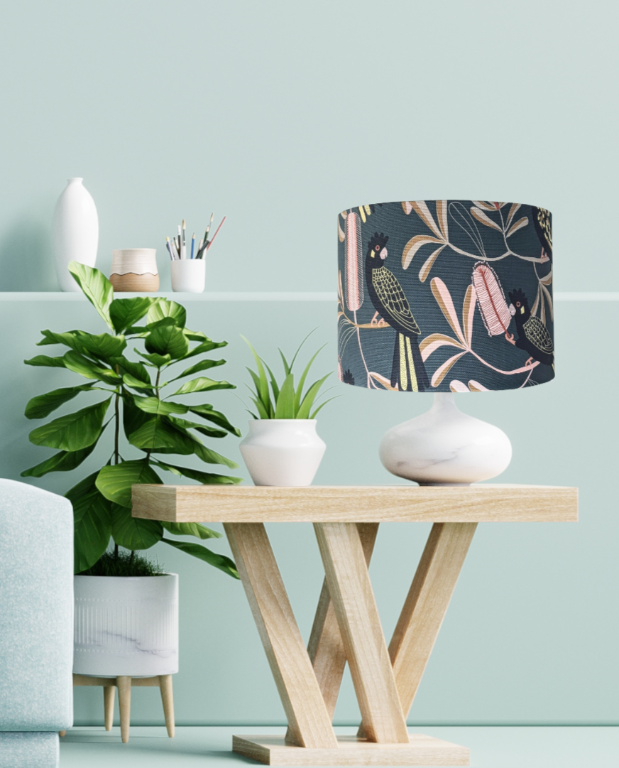 A modern wooden side table with a marble-based lamp featuring the Emotive Collection Yellow-Tailed Black Cockatoo Lampshade sits beside potted plants and art supplies against a light green wall.