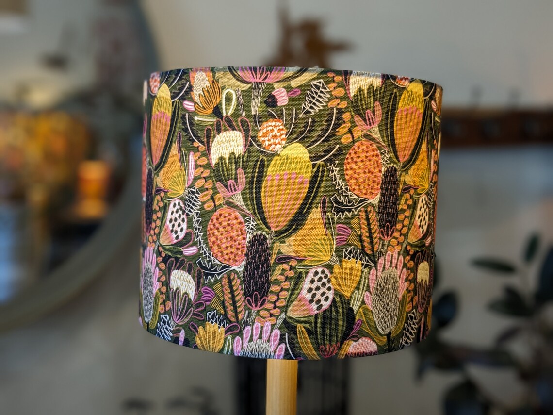 Banksia flowers on a rich green background decorate this Handmade Bush Banksia lampshade, The light has been turned on to showcase the shade when the light is switched on, a stunning design by Australian artist Jocelyn Proust. Expertly crafted in Australi