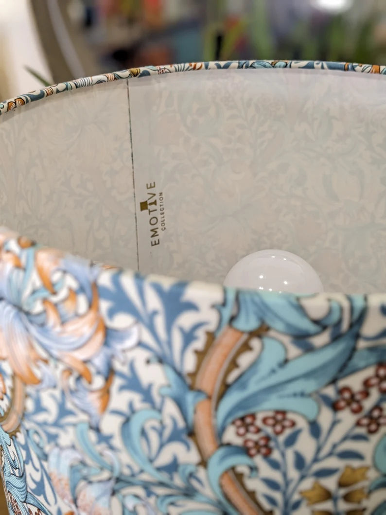 Close-up of the William Morris Golden Lily Lampshade by Emotive Collection, featuring an ornate floral design in blue, tan, and red. The inner side displays the Emotive Collection brand with a partially visible light bulb.