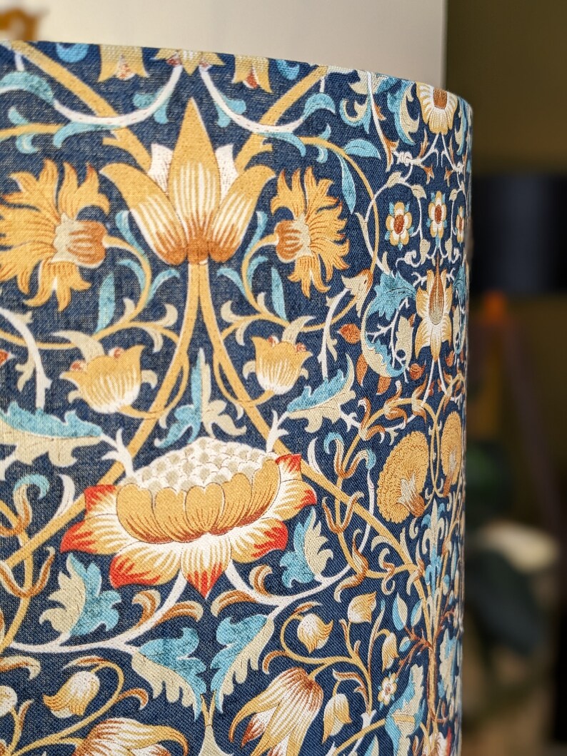 A close-up of the Emotive Collections William Morris Lodden Lampshade, showcasing an intricate blue, gold, white, and orange floral design inspired by vintage Lodden patterns. The background reveals a softly blurred elegant interior.