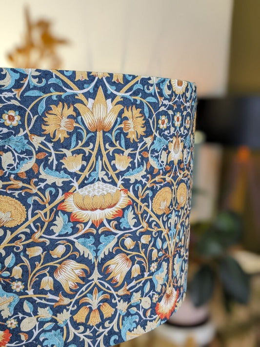 Close-up of the William Morris Lodden Lampshade by Emotive Collection, featuring a handcrafted floral pattern in blue, yellow, white, and orange tones, paired with indoor plants and soft lighting for artistic elegance.