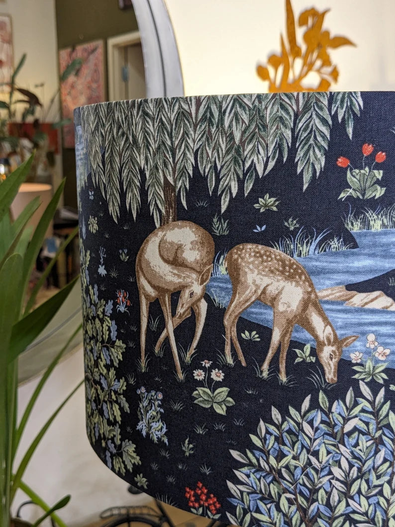 The William Morris The Brook Lampshade by Emotive Collection features a detailed illustration of two deer by a stream amid flowers, grass, and leafy trees, with softly blurred indoor plants and artwork enhancing its charming design.