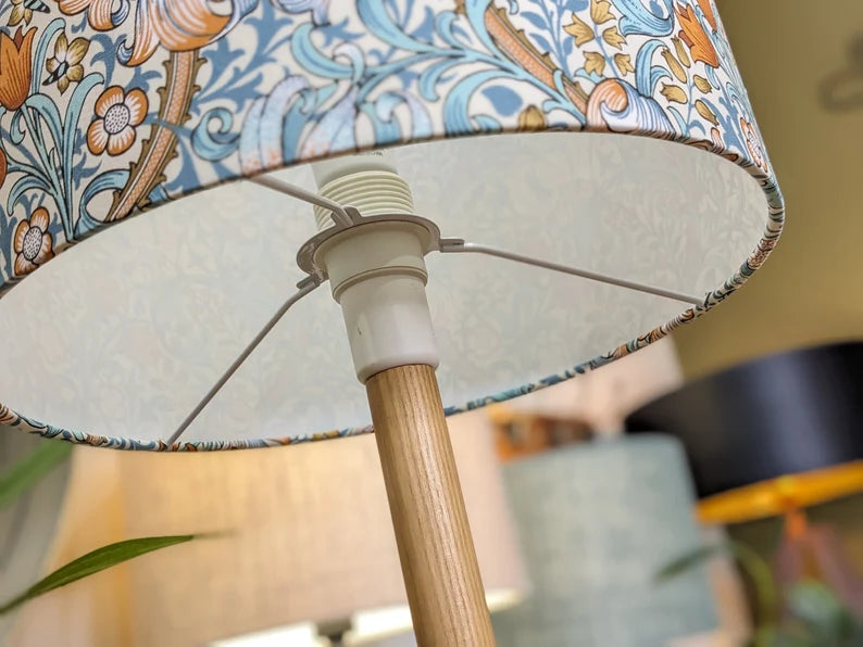Close-up of the Emotive Collection’s William Morris Golden Lily Lampshade, featuring a blue and orange floral pattern, on a wooden base. Other lamps softly blurred in the background add artistic elegance.