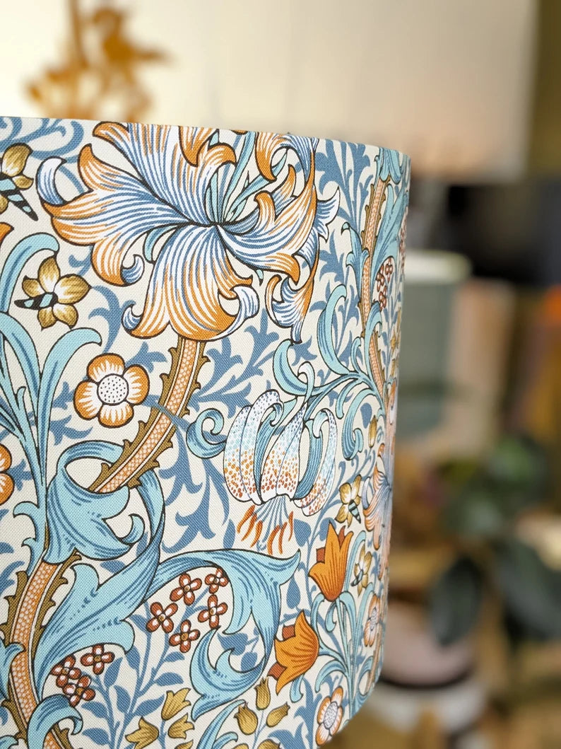 A close-up of the Emotive Collections William Morris Golden Lily Lampshade showcases its detailed blue, orange, and cream floral pattern, highlighting the artistic elegance inspired by Morris’s iconic design against a blurred backdrop.