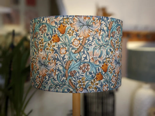 A close-up of the Emotive Collections William Morris Golden Lily Lampshade, featuring a vintage floral pattern in blue, orange, and cream with detailed swirling leaves and flowers; the background is softly blurred.