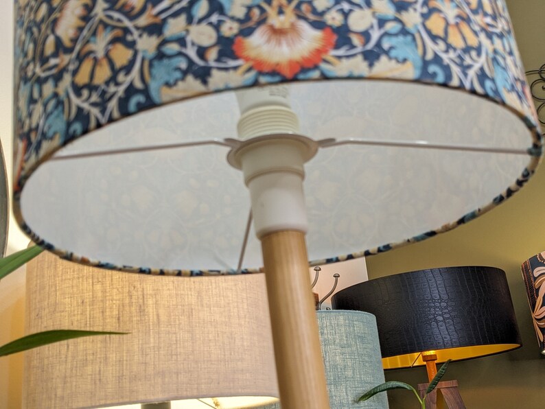 A close-up of the Emotive Collection William Morris Lodden Lampshade, showcasing its handcrafted floral design and vivid colors on a wooden floor lamp stand, with other uniquely textured and colored lamps visible in the background.