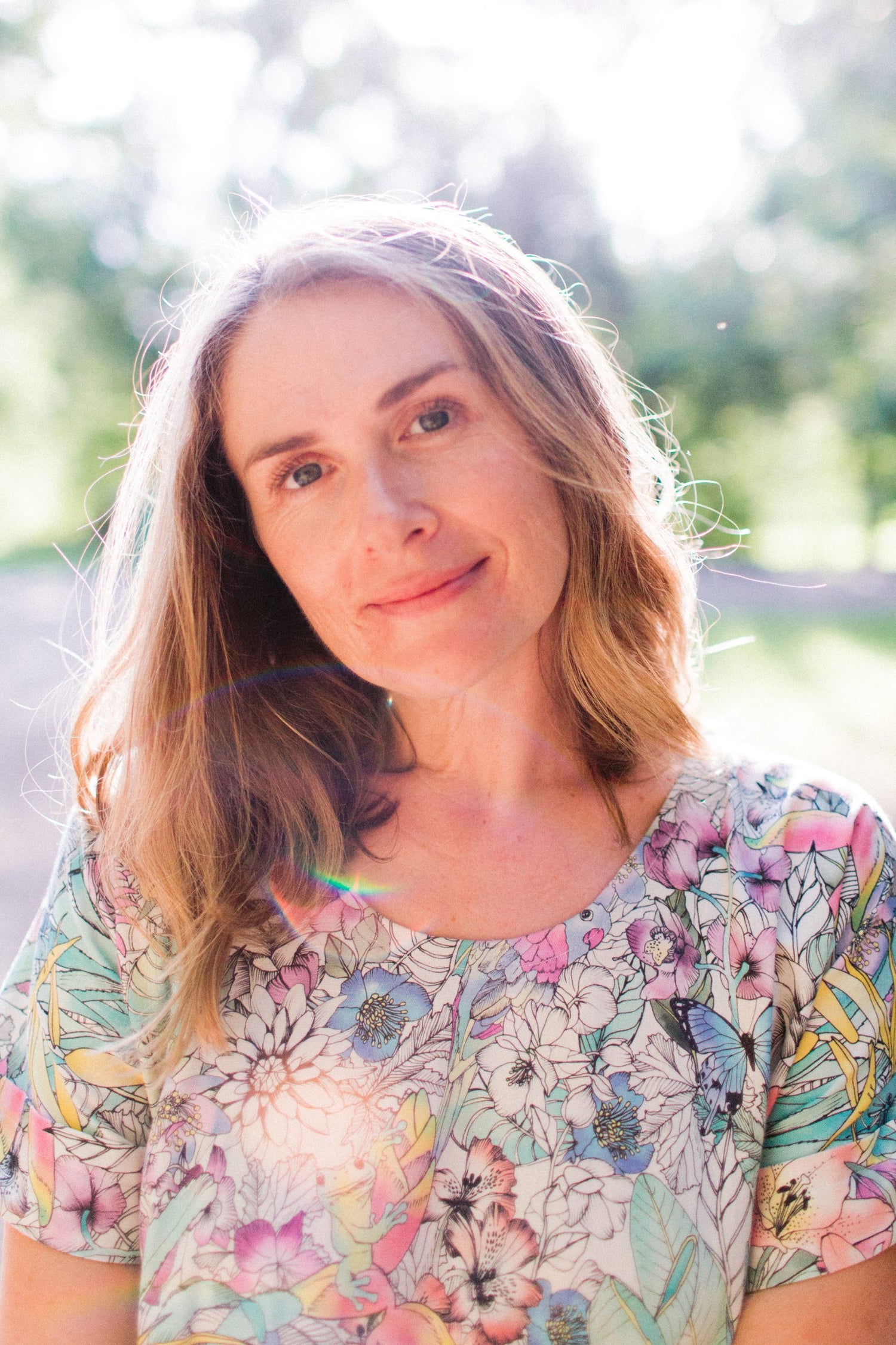 A woman with light brown hair and a floral top smiles gently at the camera, standing outdoors in sunlight with a blurred green background.
