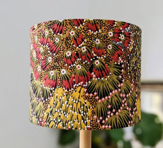 The Bush Banana Lampshade by artist Donna Abbot showcases a vibrant Aboriginal design with red, yellow, green, and white abstract shapes and dots resembling botanical motifs on a neutral background.