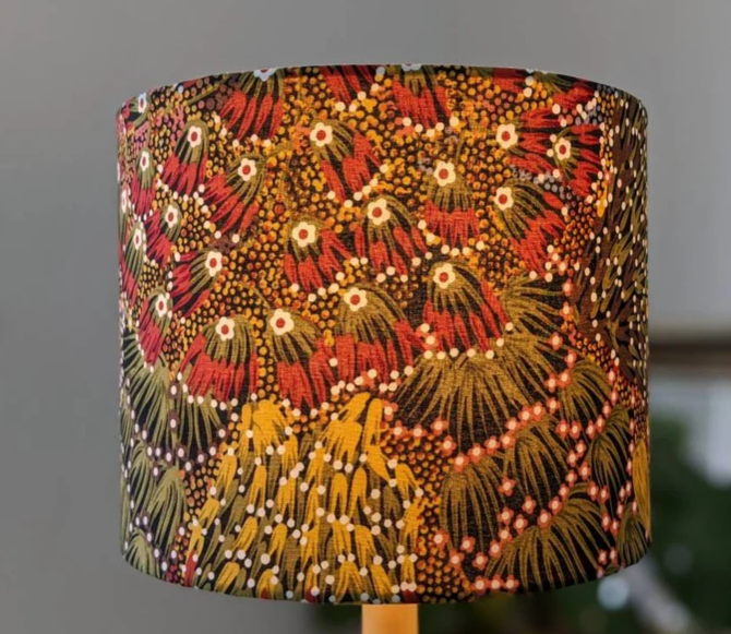 The Bush Banana Lampshade by artist Donna Abbot features vibrant fabric with abstract dots in red, yellow, green, and white, inspired by Indigenous art, standing out against a blurred neutral background.