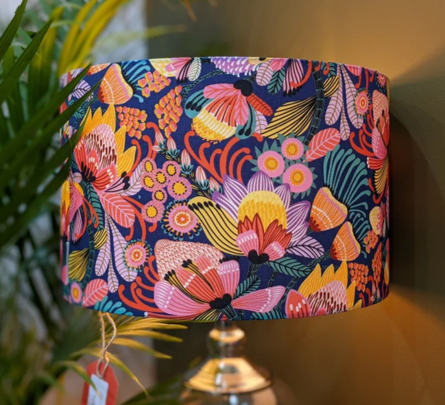 The Emotive Collections Wild Protea Blue Lampshade, handmade by Kirsten Katz with a vibrant floral design, stands on a metallic base against a warm beige wall and lush green leafy plants.