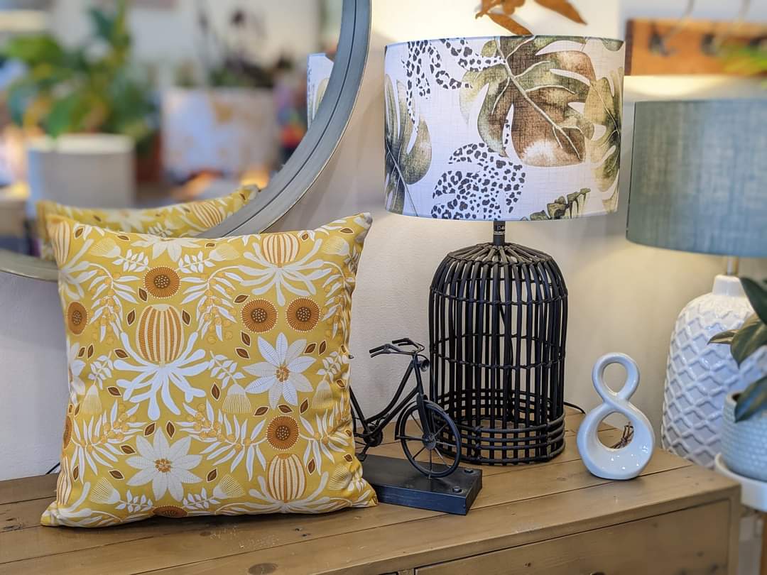 A decorative scene featuring an Emotive Collection Wattle Mustard Cushion Cover, a black wire lamp with leafy shade, a model bicycle, a white ceramic figure eight, and plants reflected in a round mirror.
