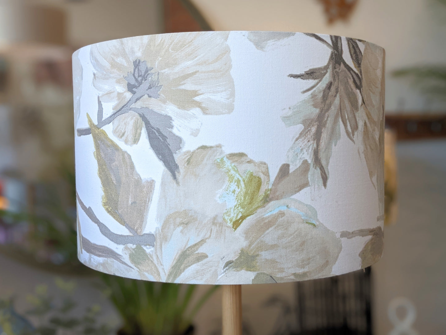 A close-up of the In Bloom Lampshade by Emotive Collection, showcasing its soft, muted floral pattern in beige, cream, green, and gray, set against a softly blurred indoor background with plants and table lamps.