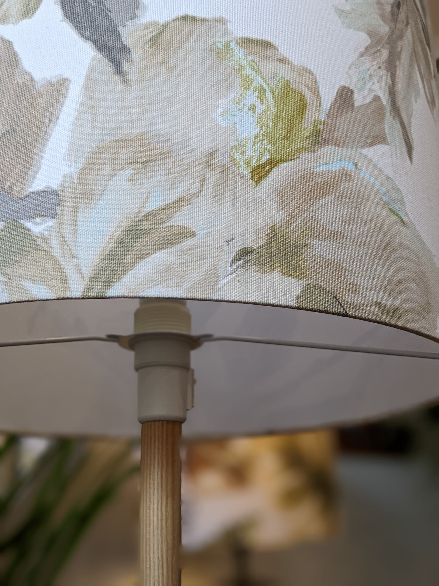 A close-up of the In Bloom Lampshade from the Emotive Collection, featuring a handmade floral pattern in soft beige, green, and light brown tones on a wooden base, set against a softly blurred background.