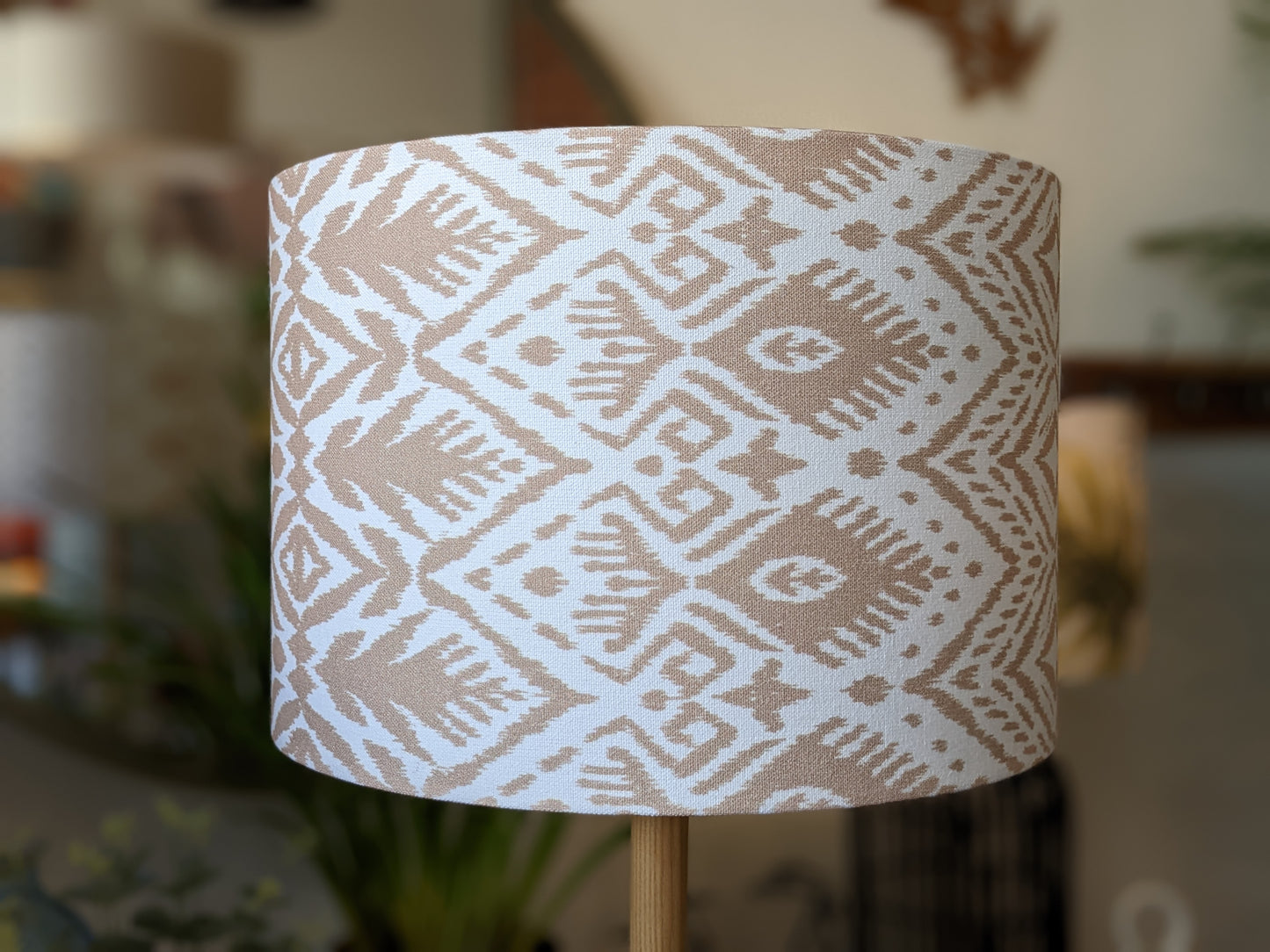 A close-up of the Ikat Lampshade from Emotive Collection shows its beige and white geometric diamond and abstract pattern. Plants and home decor appear blurred in the background, complementing this handmade piece.