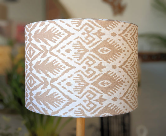 The Ikat Lampshade by Emotive Collection, featuring a beige and white geometric tribal pattern, sits on a wooden base with a softly blurred background showing touches of greenery and indoor decor.