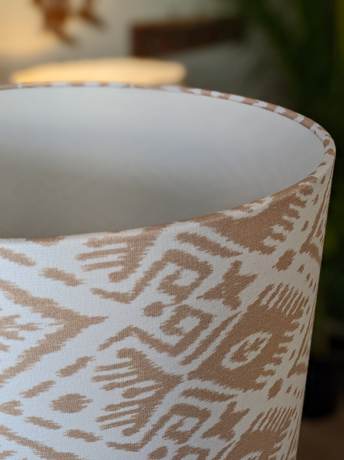 Close-up of the Ikat Lampshade from the Emotive Collection, featuring a beige and white handmade geometric tribal pattern, with a blurred background showing another lamp, a leafy green plant, and lampshades in various sizes.