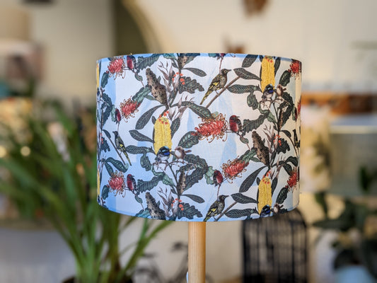 The Yellow Banksia & Native Birds Lampshade from the Emotive Collection features a handmade cylindrical design with colorful botanical and yellow bird motifs, shown indoors with blurred green plants in the background.