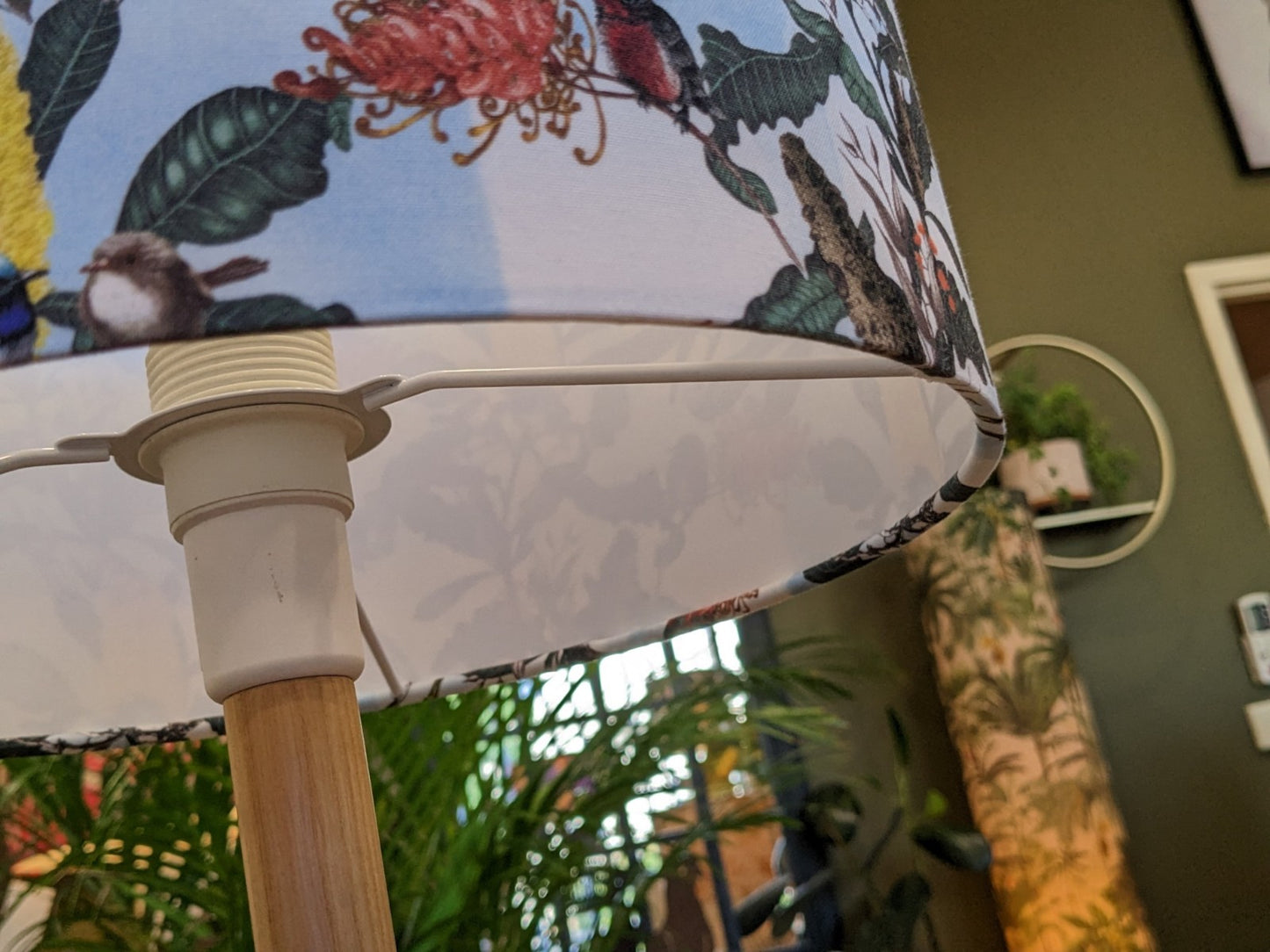 A close-up of the Yellow Banksia & Native Birds Lampshade from the Emotive Collection, featuring a vibrant botanical and bird design, set indoors with plants and a green wall in the bright background.