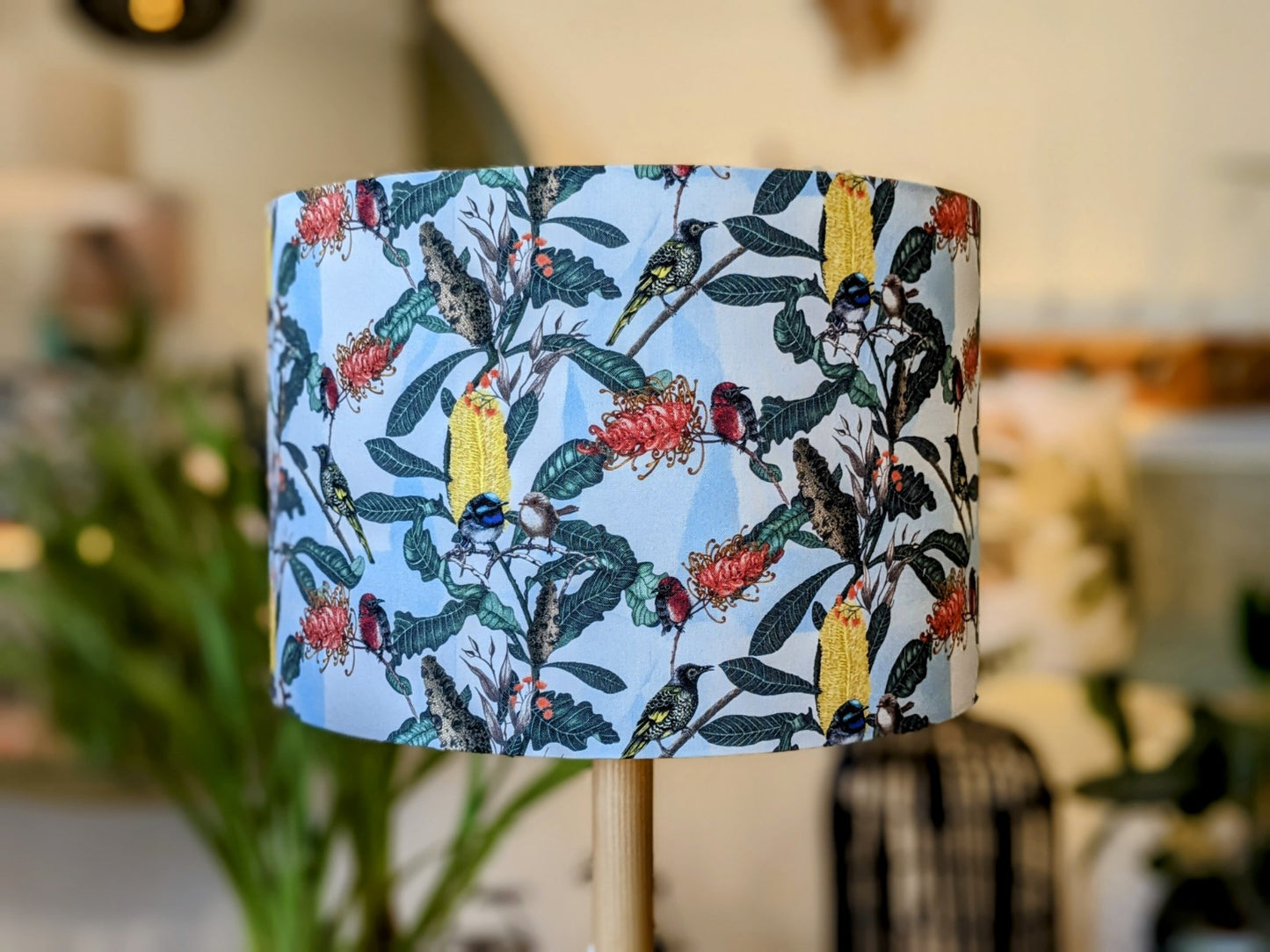 The Emotive Collections Yellow Banksia & Native Birds Lampshade is a cylindrical, handmade piece featuring Tamara Schneider’s colorful design with yellow parrots and green foliage on a light background, adding cozy ambiance to any indoor space.