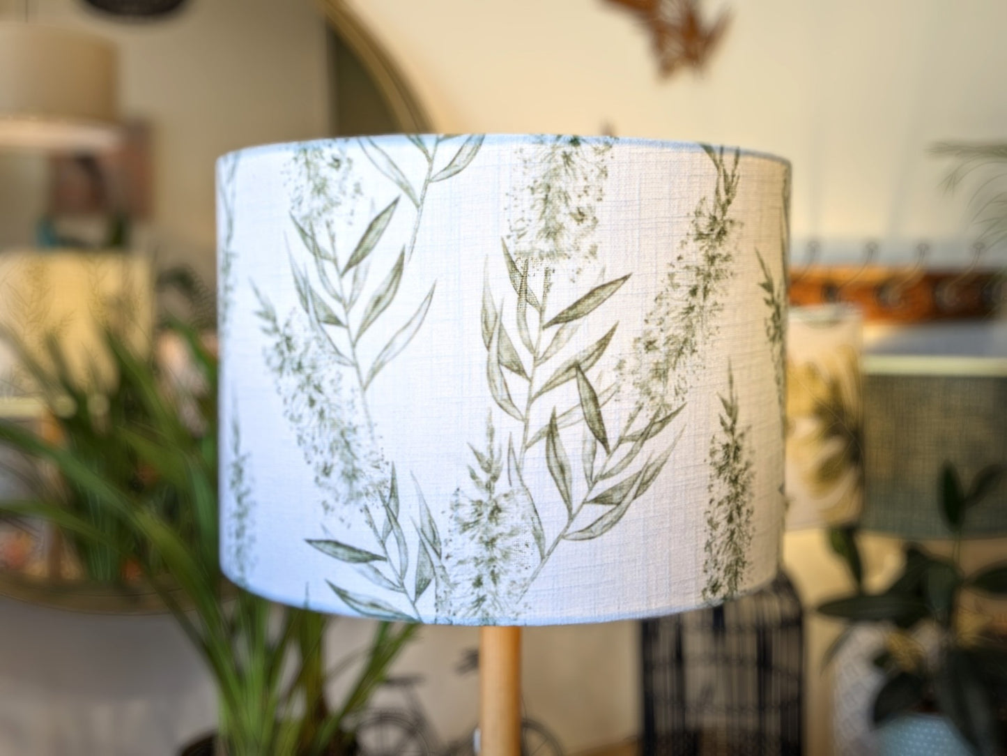 A close-up of the Emotive Collection’s Bottlebrush Green Lampshade, crafted with white linen-blend fabric and a green leafy botanical pattern, set in a bright room with softly blurred plants and decor in the background.