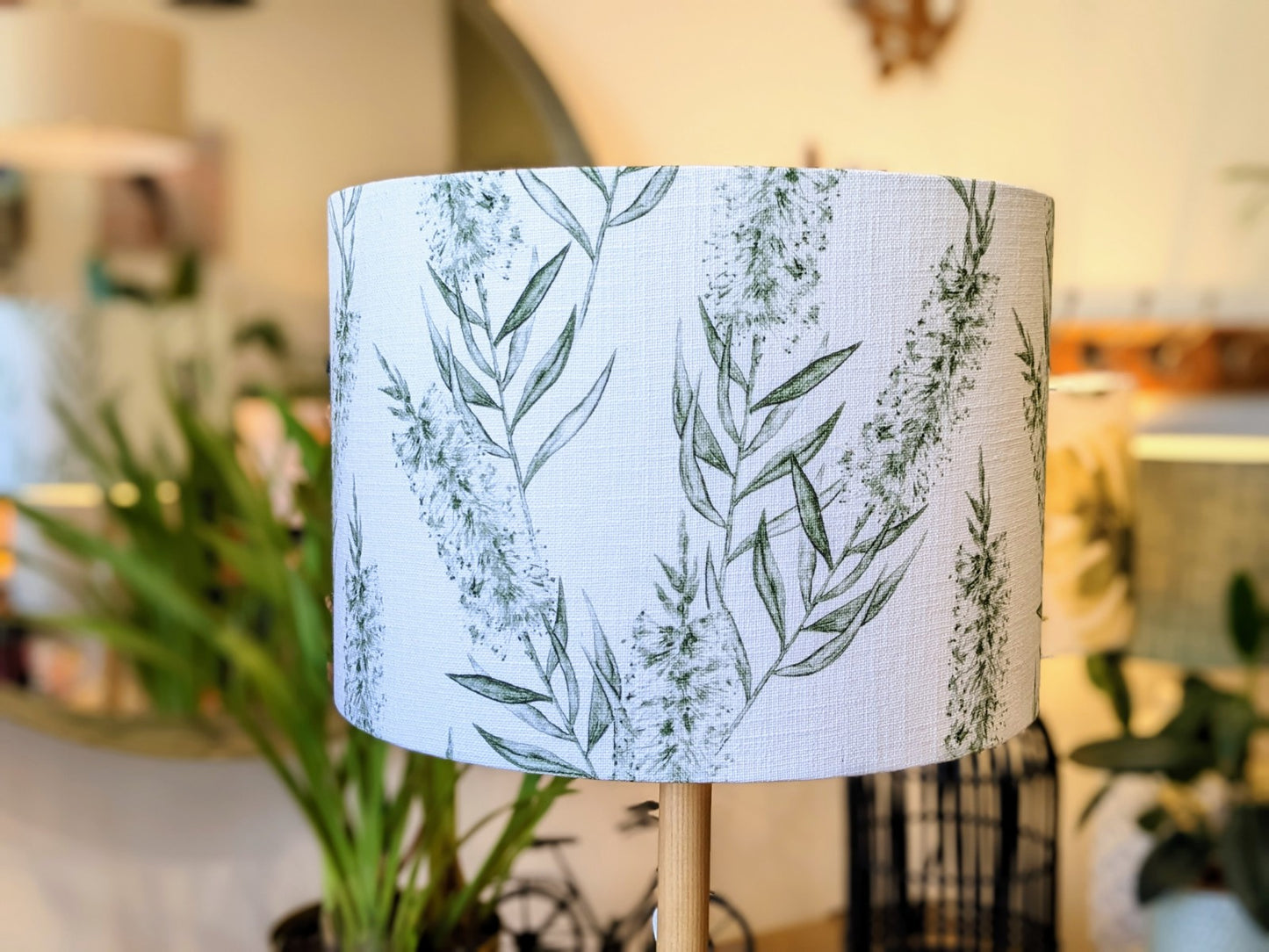 A close-up of the Bottlebrush Green Lampshade by Emotive Collection, handmade in a linen blend with a white background and green botanical leaf pattern, set in a softly blurred indoor space with plants and decor.