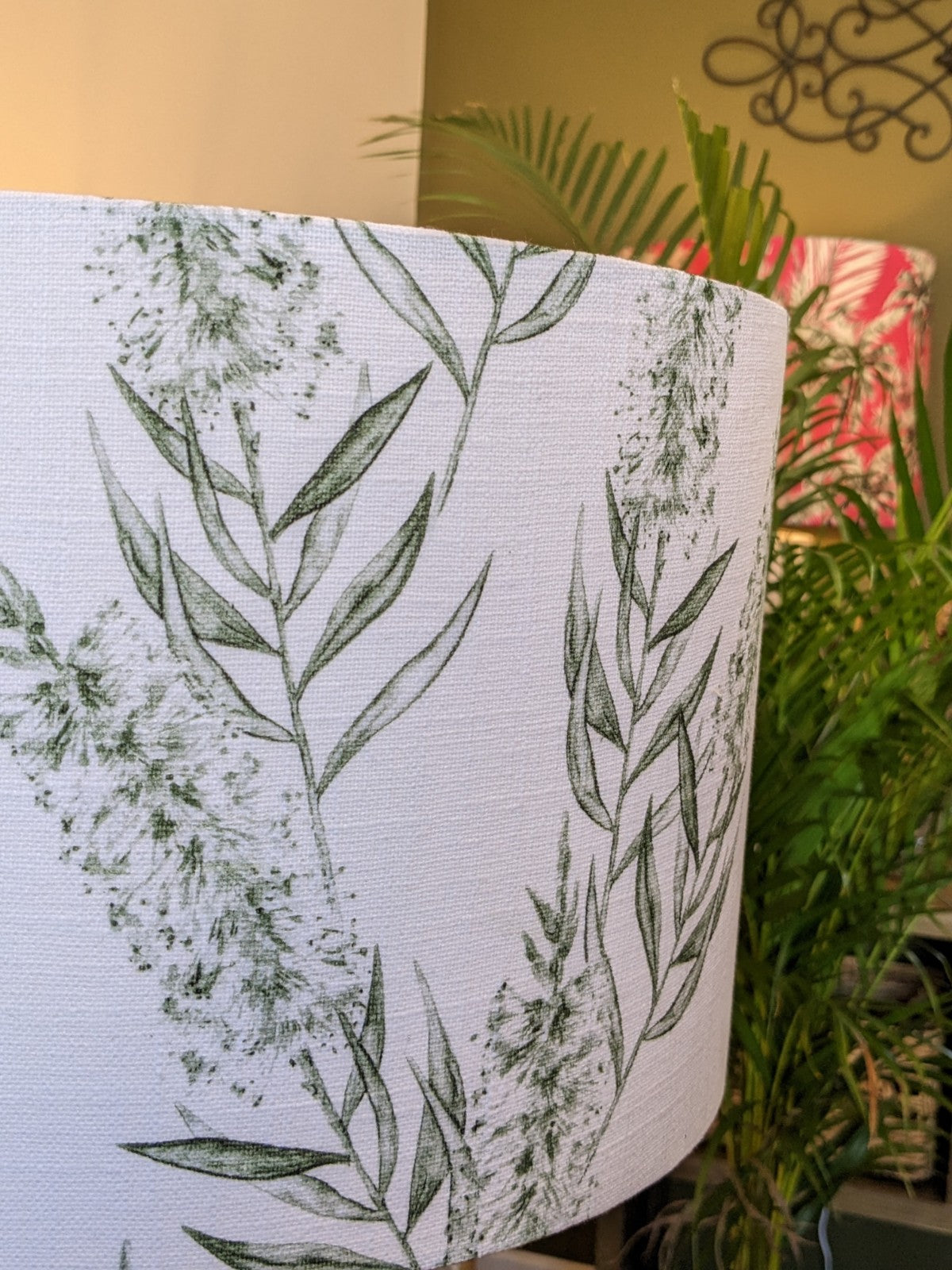 A close-up of the Bottlebrush Green Lampshade from the Emotive Collection, handmade from a linen blend with green leafy prints; lush plants and Emotive Collection wall art accent a soothing green wall in the background.