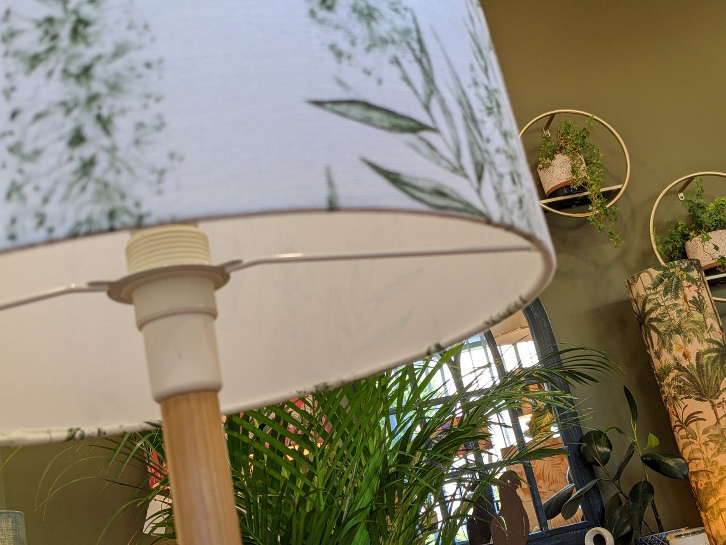 A close-up of the Bottlebrush Green Lampshade by Emotive Collection, featuring a white leafy-patterned linen blend, set against green walls with lush plants and wall-mounted planters in a cozy, well-lit room.