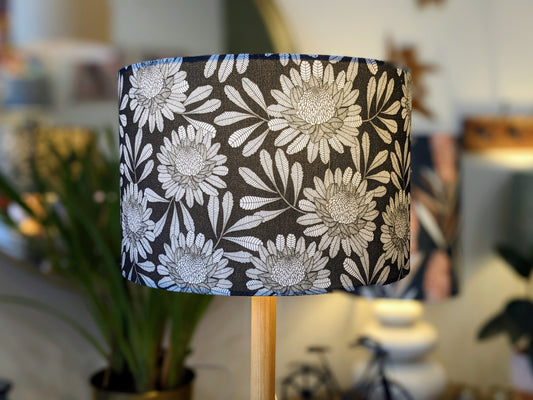 A close-up of the Emotive Collection’s Australian Waratah Lampshade featuring a white floral pattern on black, inspired by Jocelyn Proust, atop a wooden base in a stylish room with soft lighting and greenery.