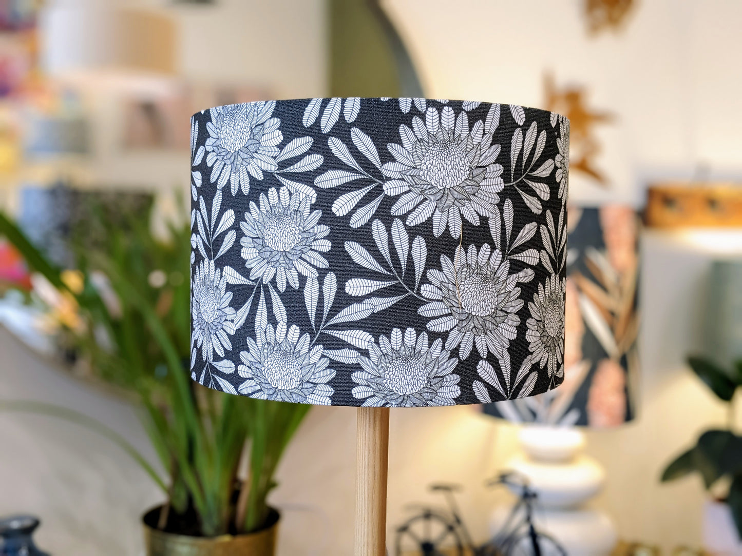 A close-up of the Emotive Collection’s Australian Waratah Lampshade shows a handmade design with large white floral details on a black base. Blurred plants and soft home decor accent the warmly lit background.