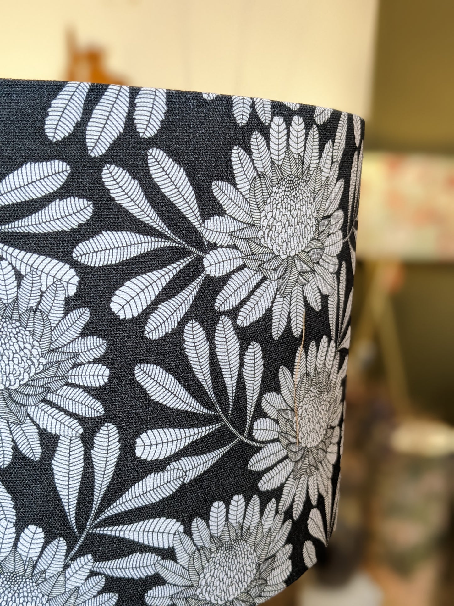 A close-up of the Australian Waratah Lampshade by Emotive Collection, handmade with a black base and white floral pattern that features detailed leaves and large, intricate flowers, set against a softly blurred background.