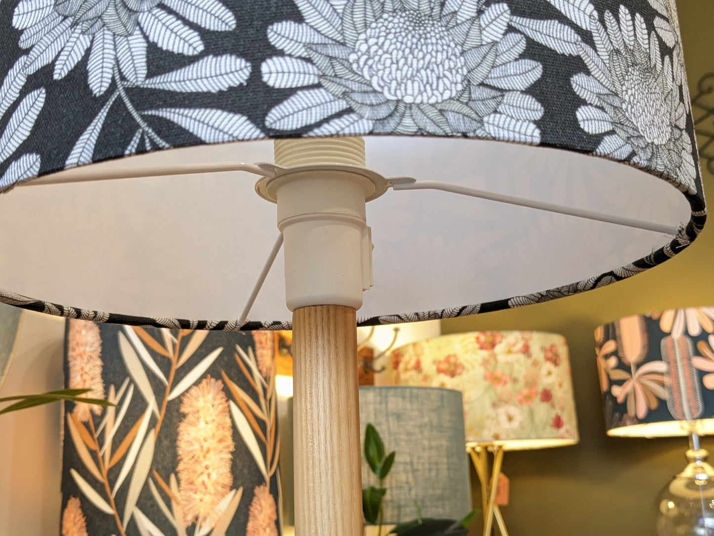 A close-up of the Emotive Collection’s Australian Waratah Lampshade shows its handmade design with a white floral pattern on a dark base, inspired by Jocelyn Proust. Other Emotive Collection lamps with abstract and floral prints blur in the background.