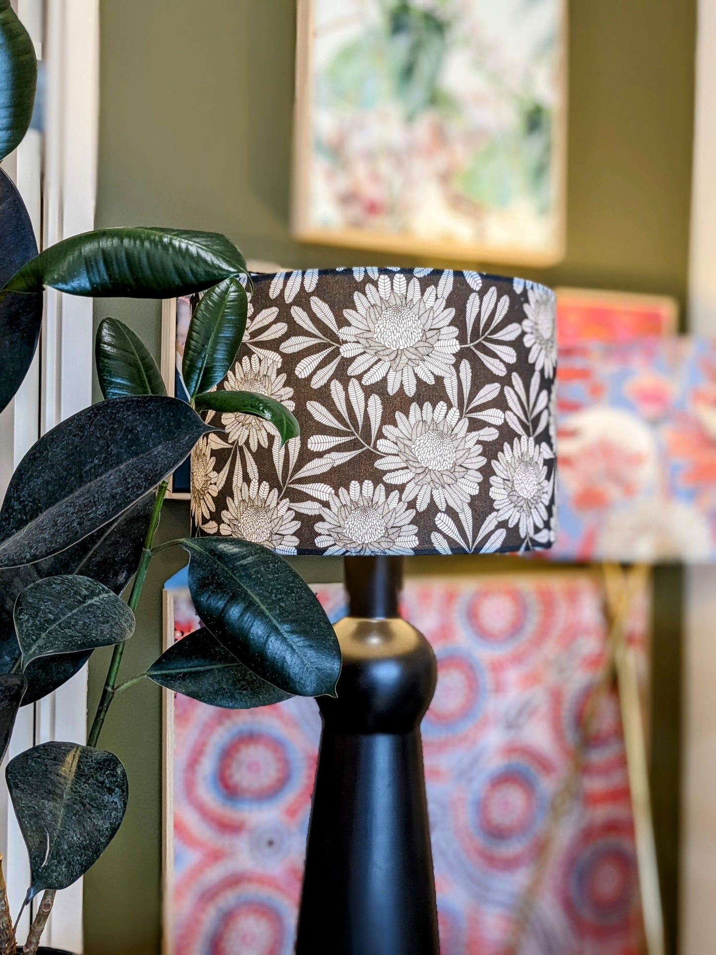An Emotive Collection Australian Waratah Lampshade with a handmade floral design in black stands beside a green-leafed plant, with vibrant abstract art by Jocelyn Proust visible in the blurred background.