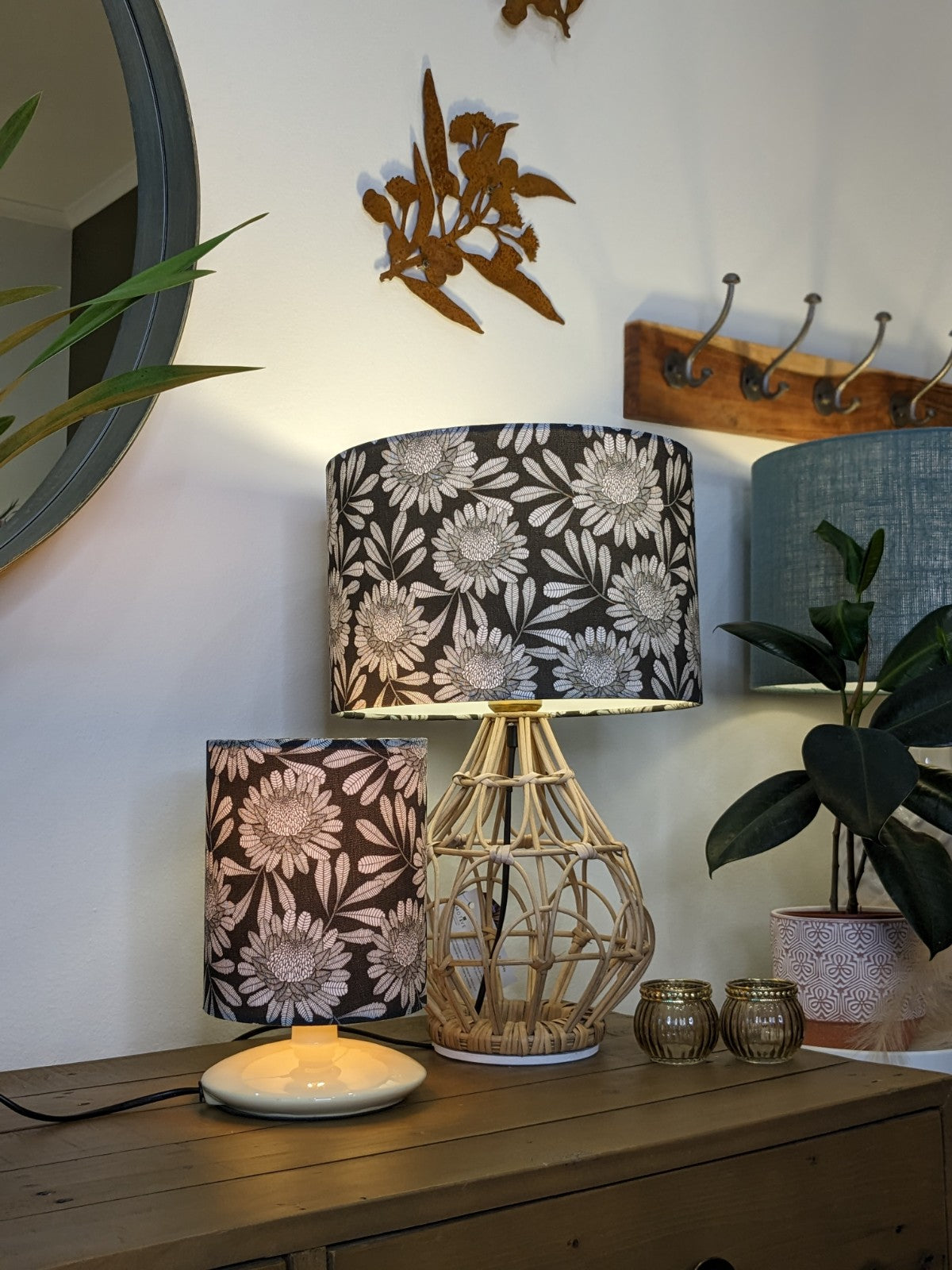 Two Emotive Collection Australian Waratah Lampshades sit on a wooden table. A decorative mirror, wall hooks, potted plant, and metal leaf wall art complete this cozy interior with eclectic touches and subtle abstract prints in the background.