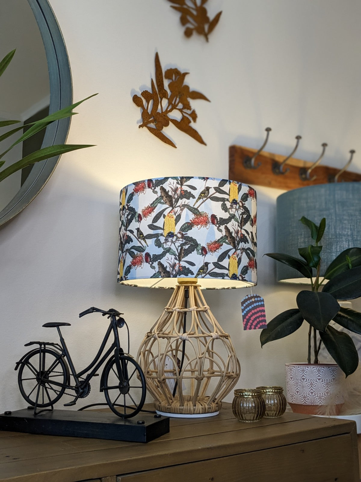 A wooden table with the Yellow Banksia & Native Birds Lampshade from the Emotive Collection, a rattan lamp, metal bicycle decor, gold candle holders, potted plant, and a round mirror on the wall.