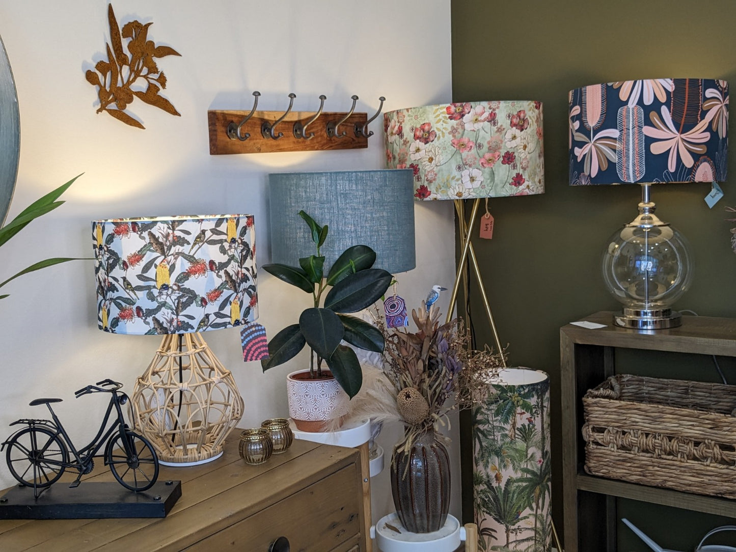 A cozy interior by Tamara Schneider features the Yellow Banksia & Native Birds Lampshade from the Emotive Collection, a potted plant, dried flowers, wicker baskets, a miniature bicycle, and a wall hook rack set against green and white walls.
