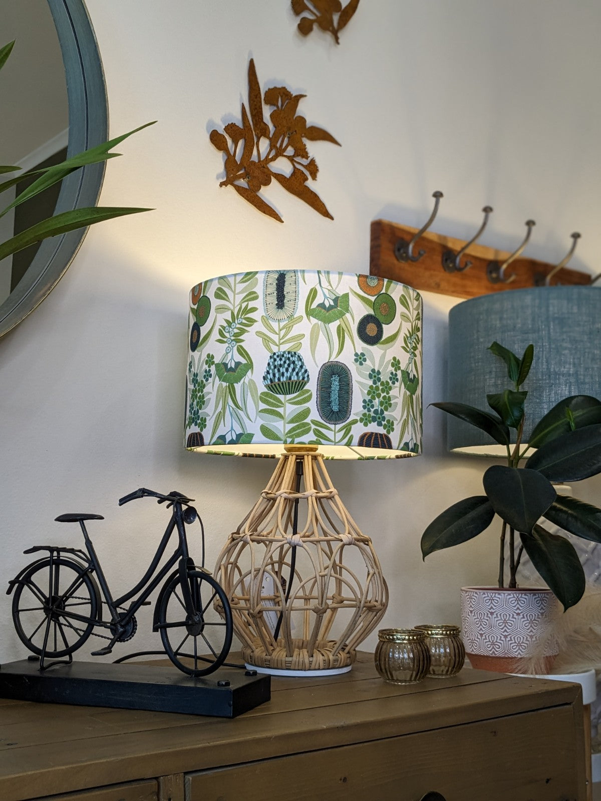 A decorative table with a rattan lamp featuring the Emotive Collection Green Australiana Lampshade, a metal bicycle figurine, two small candle holders, a potted plant, wall hooks, and bird-shaped wall art.
