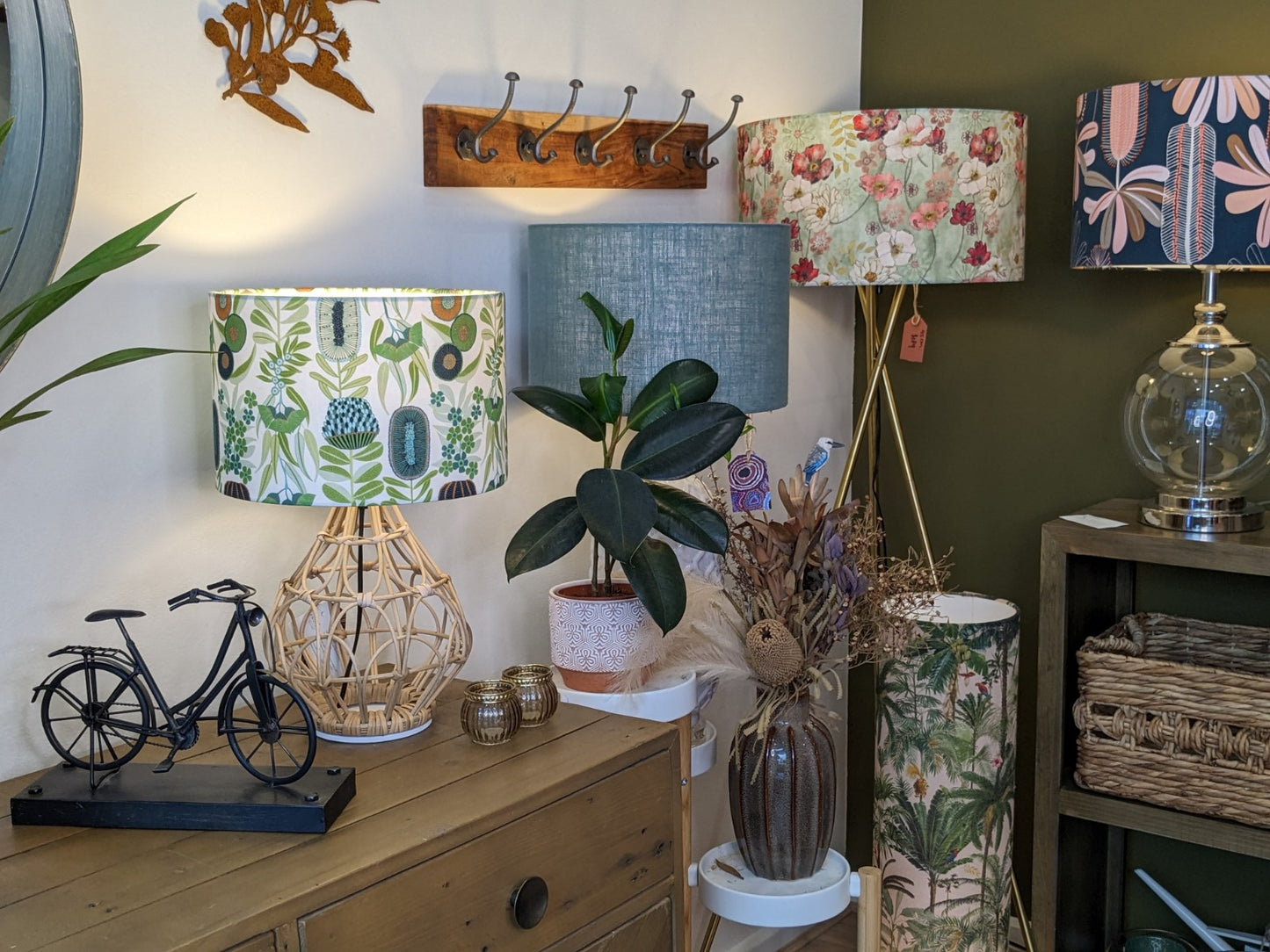 A cozy room corner with a wooden table, Emotive Collections Green Australiana Lampshade, patterned lampshades, a small plant, woven baskets, a decorative bicycle, and dried flowers in a vase against white and green walls.