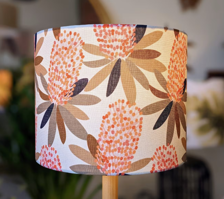 The Coastal Banksia Lampshade by Emotive Collection features stylized pink and orange flowers with brown and green leaves, inspired by Jocelyn Proust designs, shown against a blurred indoor background.