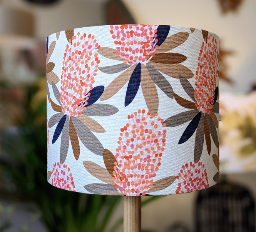 The Coastal Banksia Lampshade by Emotive Collection is a handmade shade with pink, orange, and brown flowers and navy blue leaves on white, inspired by Jocelyn Proust designs and set on a modern wooden stand.