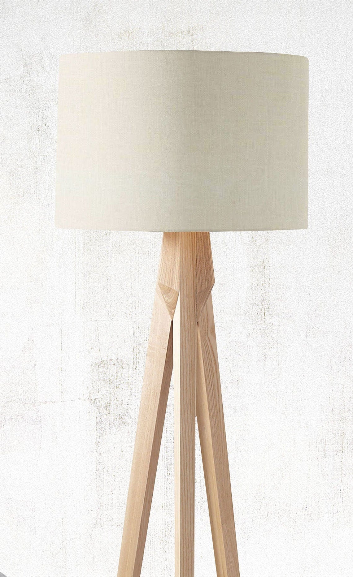 The Emotive Collection Cream Premium Linen Lampshade sits atop a modern tripod floor lamp with a light wooden base, ideal for natural interiors and set against a textured, light-colored wall.
