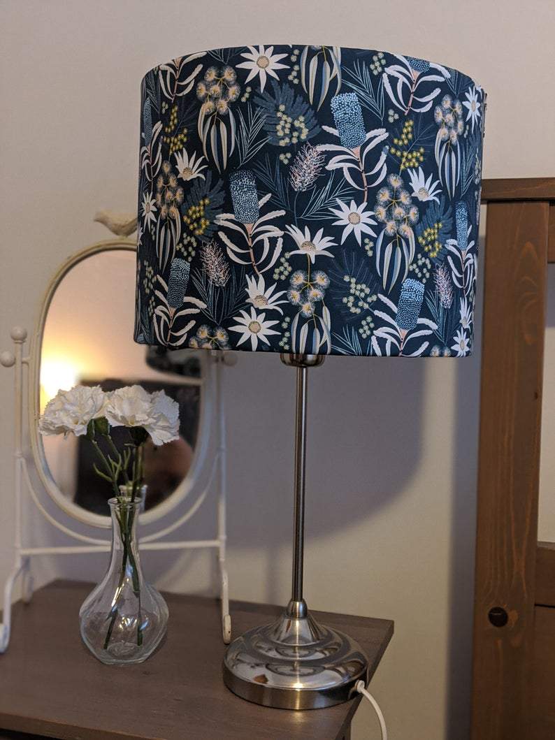 A bedside table with a tall silver lamp featuring Emotive Collection’s Blue Native Floral Lampshade, a clear vase of white flowers, and a round mirror in the background.