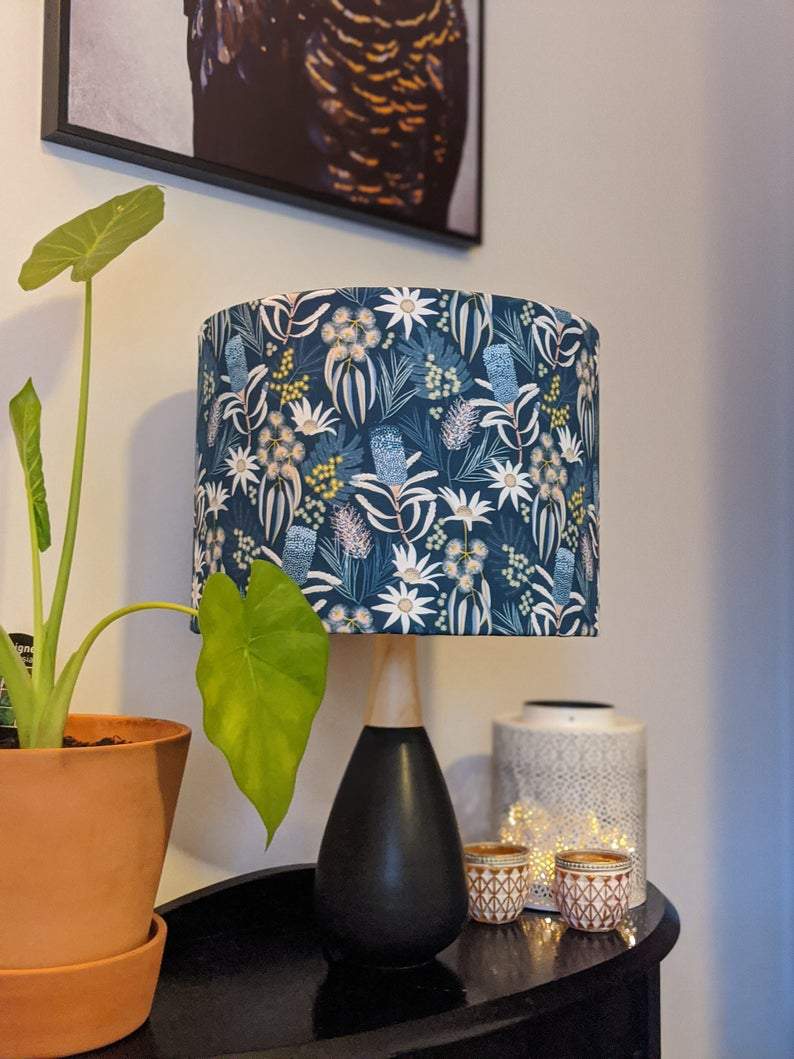 A Blue Native Floral Lampshade from the Emotive Collection sits on a dark table next to a potted plant with large green leaves, decorative candles, and a white textured container. A framed picture hangs above.