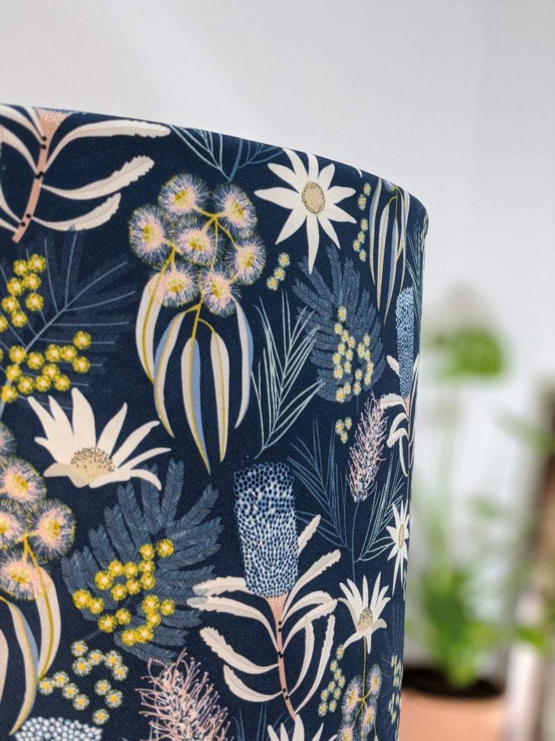 Close-up of the Blue Native Floral Lampshade by Emotive Collection, featuring a soft yellow, white, and green botanical pattern on dark blue; a blurred plant appears in the background.