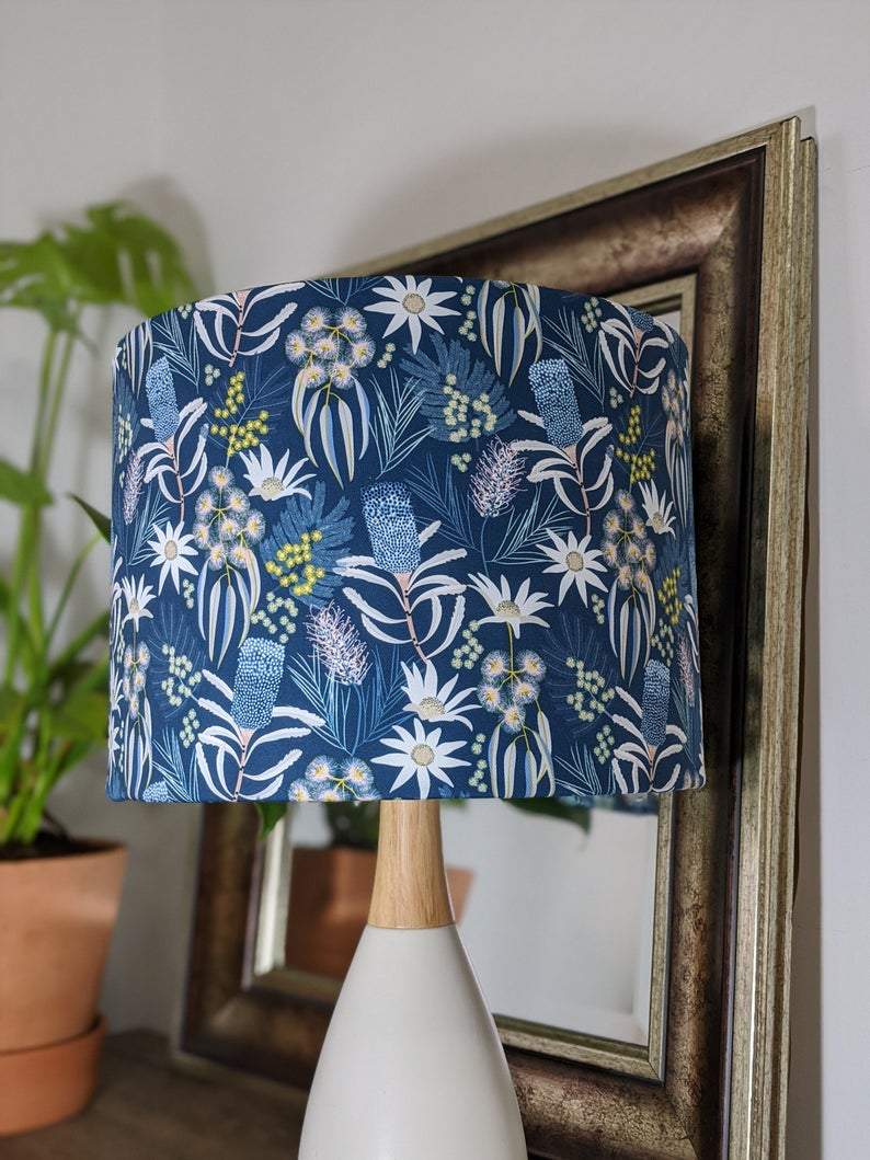 The Emotive Collection Blue Native Floral Lampshade, featuring a botanical pattern by Jocelyn Proust, sits on a white and wooden base beside a framed mirror and potted plant.