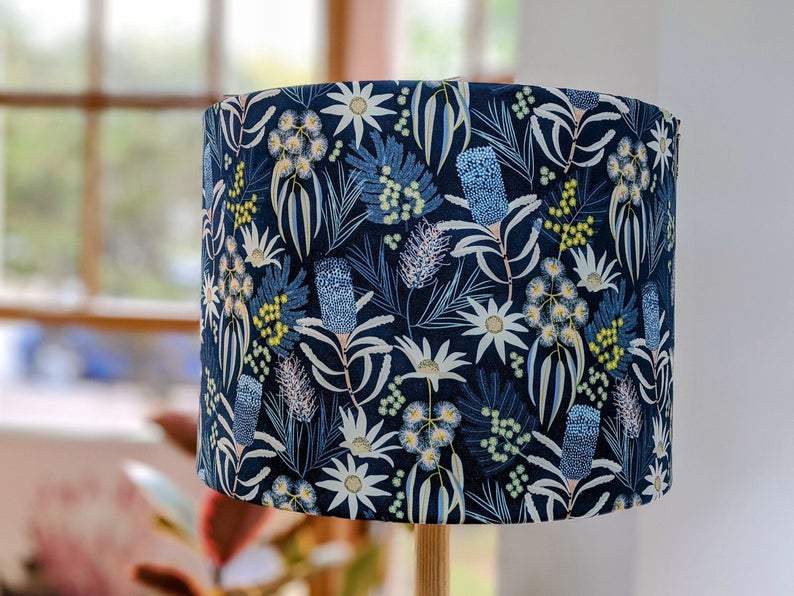 The Emotive Collections Blue Native Floral Lampshade features a vibrant botanical pattern with blue, white, and yellow flowers and leaves on a dark background, designed by Jocelyn Proust. It stands indoors near a window with blurred greenery outside.